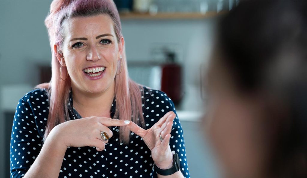 woman with pink hair and spotty shirt signing to person in the foreground