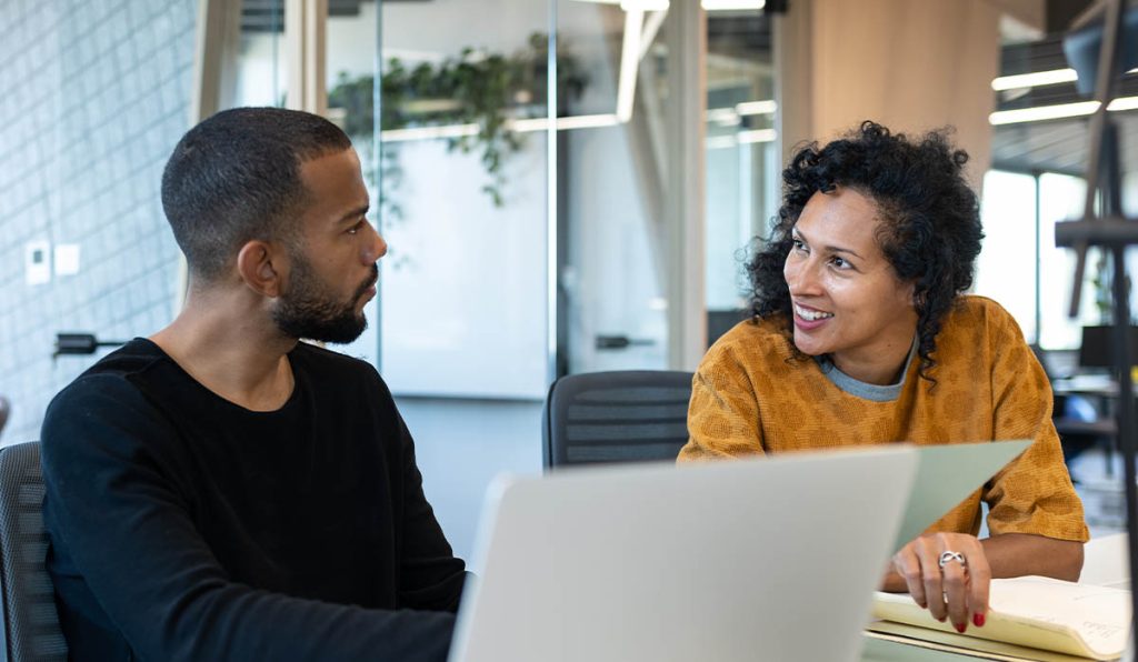 Two people having a conversation at a desk in front of an open laptop.